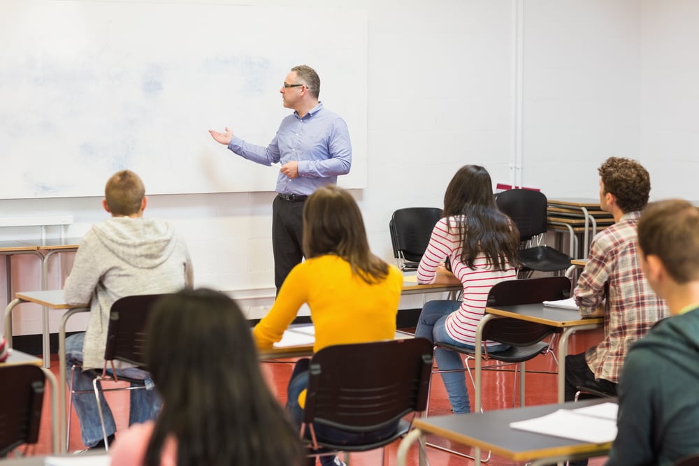 Rear view of students attentively listening to male teacher in the classroom